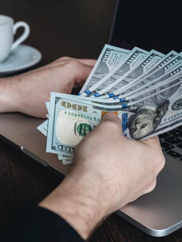 a person at a table counting u.s. dollars near an open laptop and a coffee cup, exploring ways to make money from their blog.