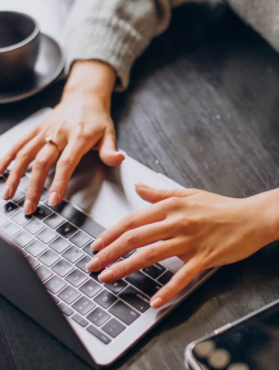 a person starting a blog by typing on a laptop keyboard with one hand, while their other hand is placed on the table next to a smartphone and coffee cup.