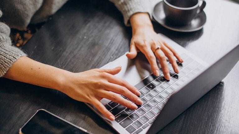 a person typing on a laptop keyboard on a wooden desk, working on a blog to make money online, with a smartphone and a cup of coffee nearby.