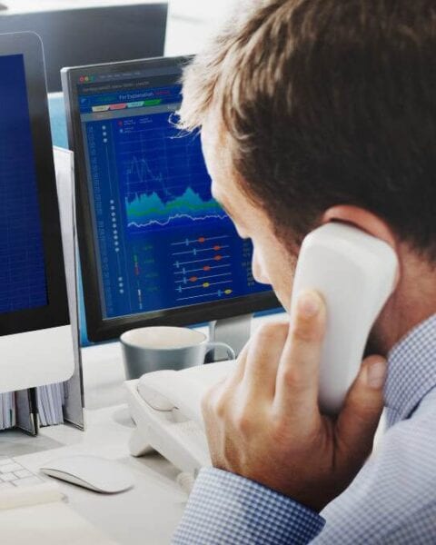 a man is talking on the phone in front of two monitors, strategizing ways to increase sales and optimize seo keywords.