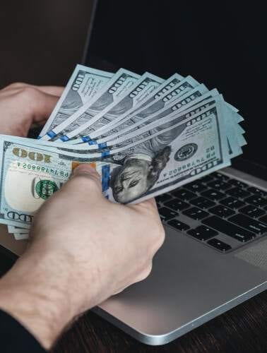 a man is holding a stack of dollar bills in front of a laptop, researching the best passive income ideas.