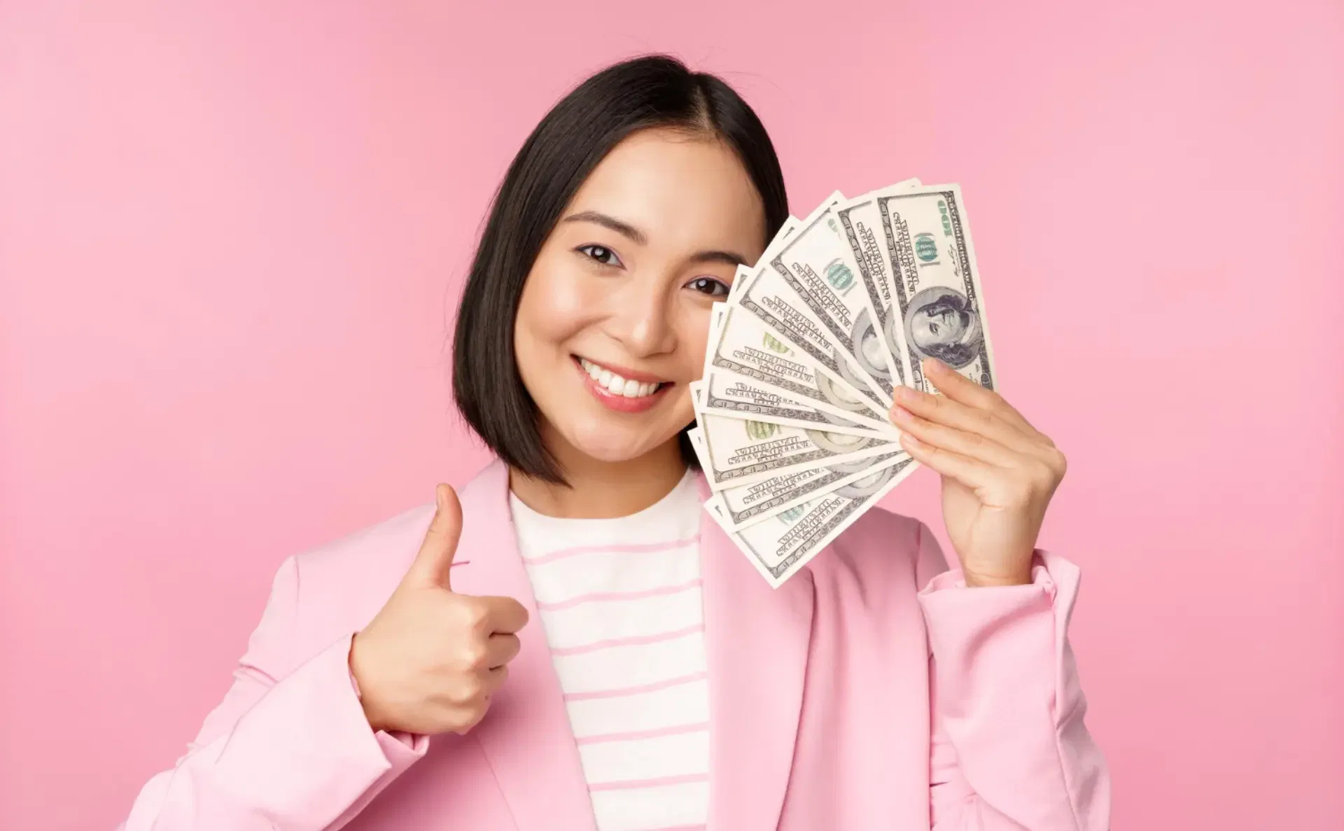 Sarah Titus in a pink blazer, a self-made millionaire she, is smiling and giving a thumbs up while holding a fan of us dollar bills against a pink background.