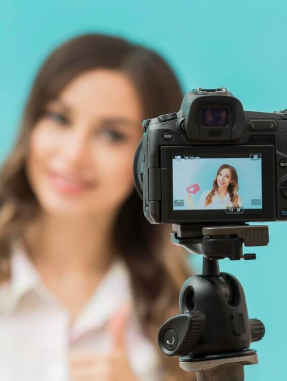 camera on a tripod focusing on a blurred young woman in the background, displayed in its screen against a teal backdrop as she offers free advice for making money.