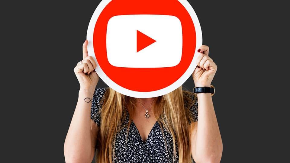 a woman holds a large youtube play button sign in front of her face against a dark background, symbolizing her journey as a full-time youtuber.