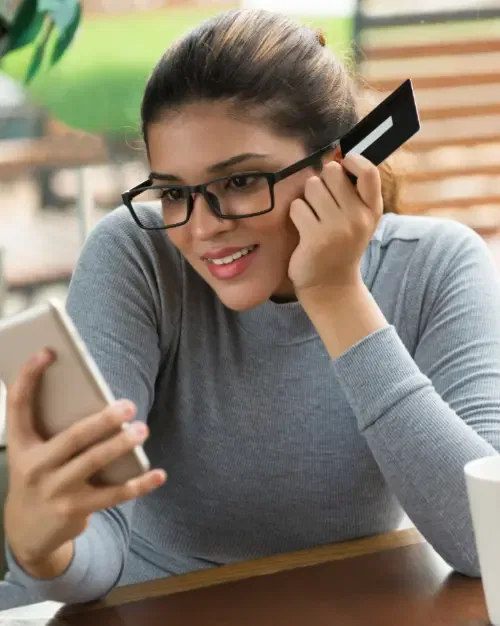 A person wearing glasses and a gray top holds a credit card to their ear while looking at a smartphone, seemingly brainstorming business ideas.
