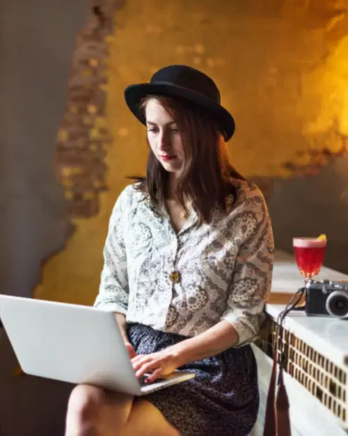A woman wearing a hat uses a laptop while sitting at a counter with an exposed brick wall, a camera, and a cocktail nearby, possibly brainstorming business ideas for a niche market.
