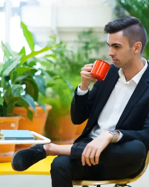 A person in a black suit sits on a chair, holding a red mug, surrounded by green potted plants indoors, likely contemplating new business ideas for tapping into a niche market.