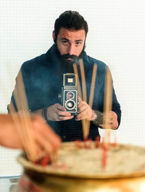 a man with a beard holding a vintage camera, focusing intently, with a blurred foreground of candles on a cake, capturing the essence of social media authenticity.