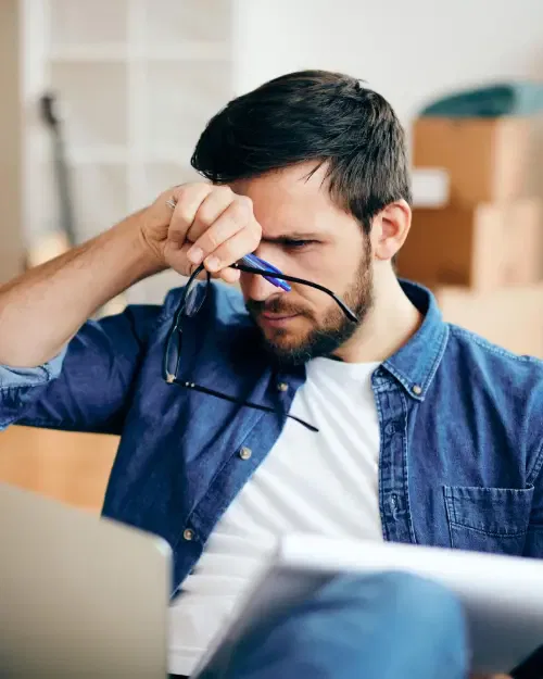 a man with a beard and casual attire holds his glasses in one hand, appearing focused or stressed, possibly contemplating unexpected challenges while sitting in front of a laptop.
