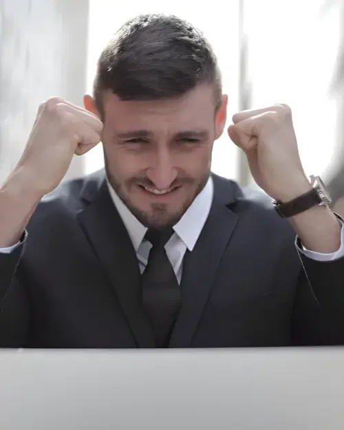 a man in a dark suit and tie clenches his fists in celebration, smiling slightly, as if recognizing the success of implemented strategies while appearing to look at something in front of him.