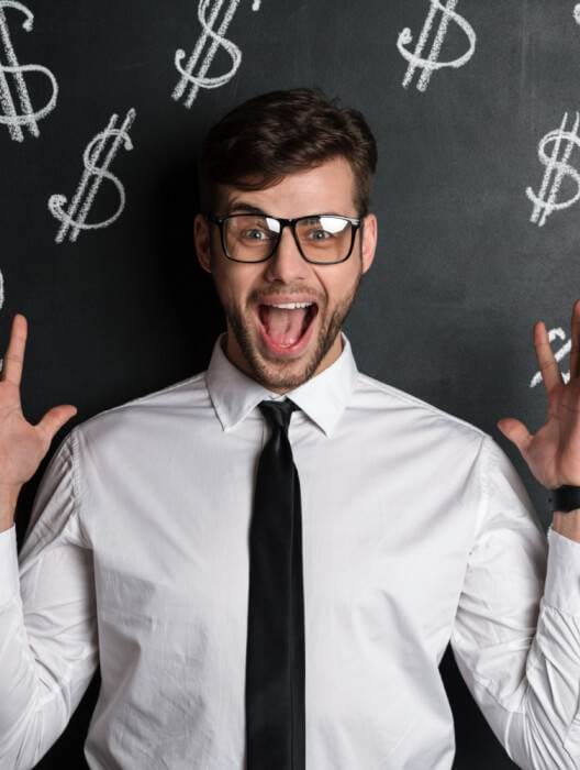 A joyful man in a white shirt and black tie, wearing glasses, excitedly raises his hands against a chalkboard background with dollar signs drawn on it, celebrating his success in doubling their online income.