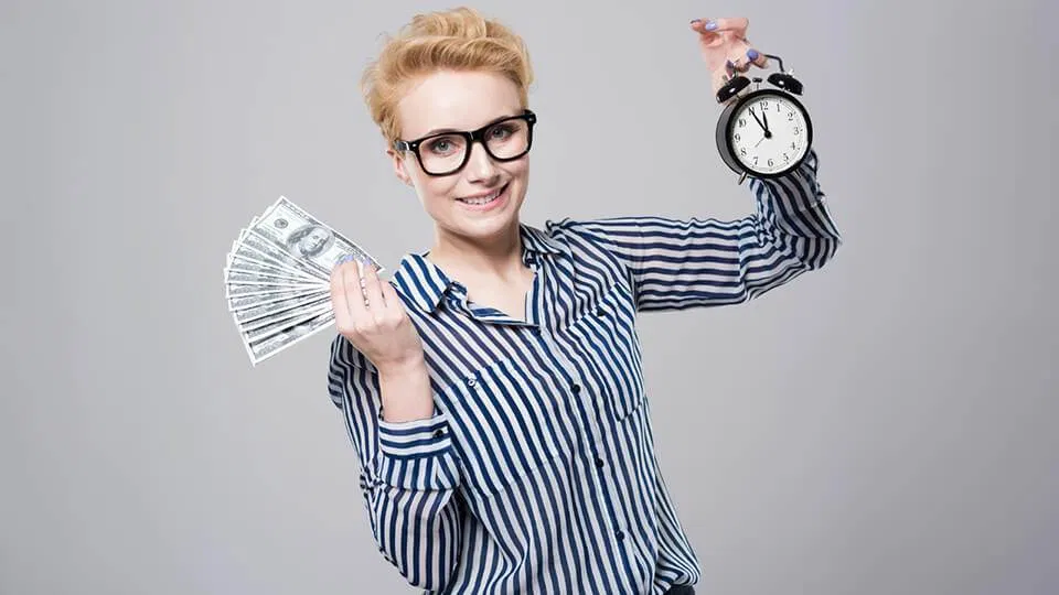 a woman with blonde hair, wearing glasses and a striped shirt, holds up a fan of extra money in one hand and an alarm clock in the other, smiling at the camera.