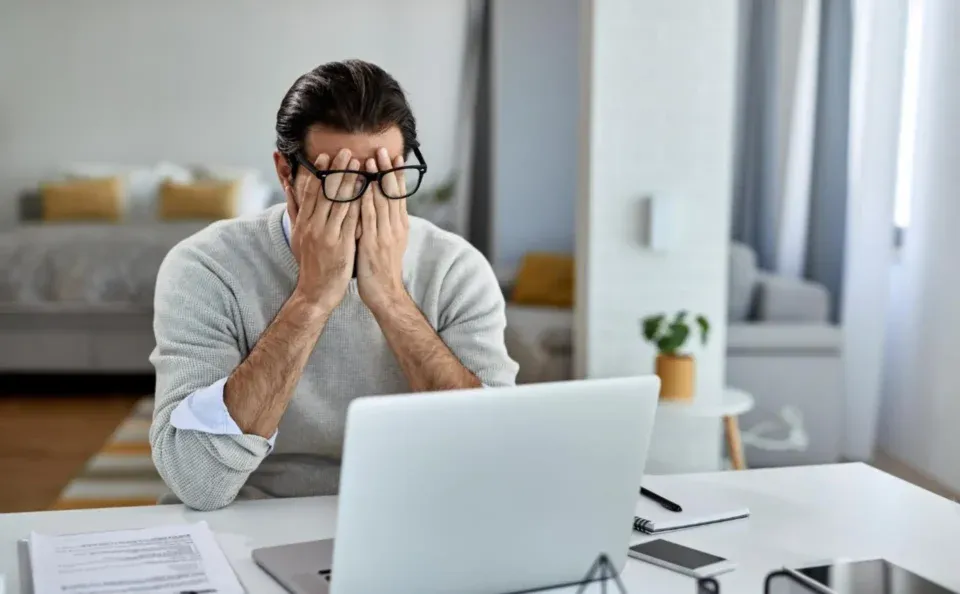 a person sits at a desk with their hands covering their face in front of a laptop, appearing stressed or tired due to unexpected challenges. a document is on the table, and a bed and plant are visible in the background.