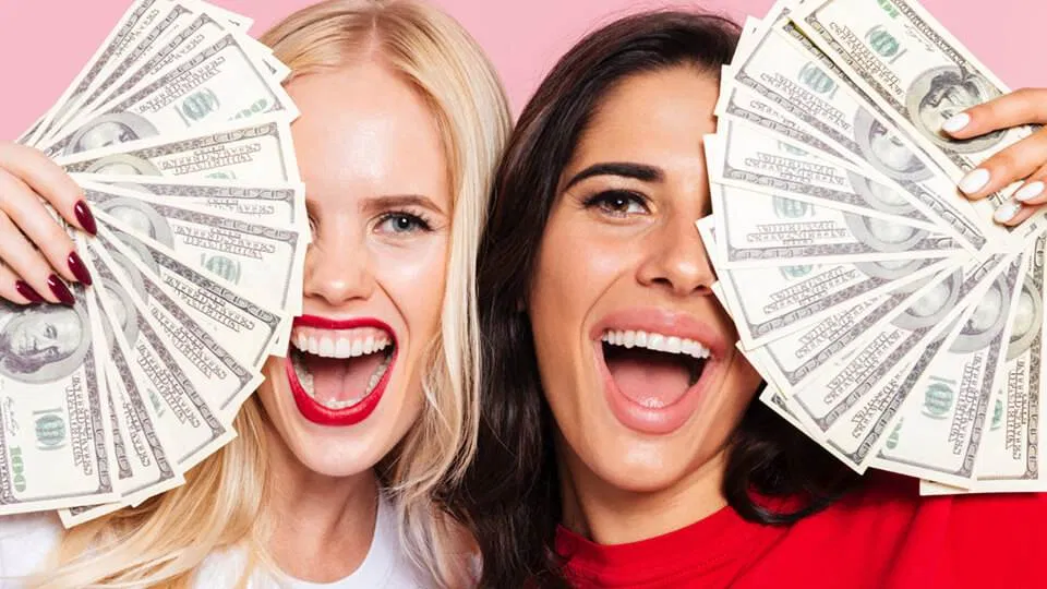 two joyful women holding multiple 100 dollar bills against a pink background, celebrating their success in doubling their online income.