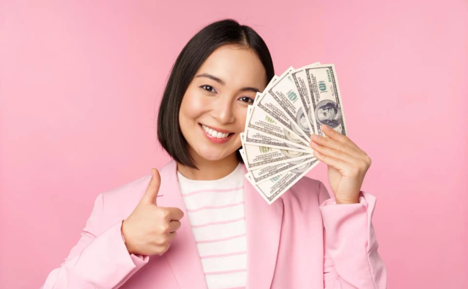 Sarah Titus in a pink blazer, a self-made millionaire she, is smiling and giving a thumbs up while holding a fan of us dollar bills against a pink background.