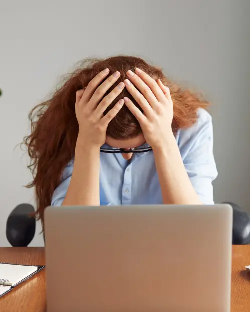 a person unexpectedly sitting at a desk with their head in their hands, in front of a laptop.