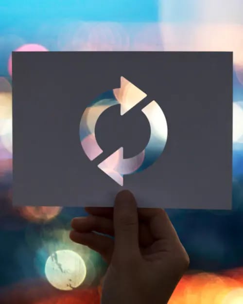 a hand holding a piece of paper with a refresh symbol cut out, revealing a blurred background of colorful lights, symbolizes the mastering strategies for creating sustainable income streams.