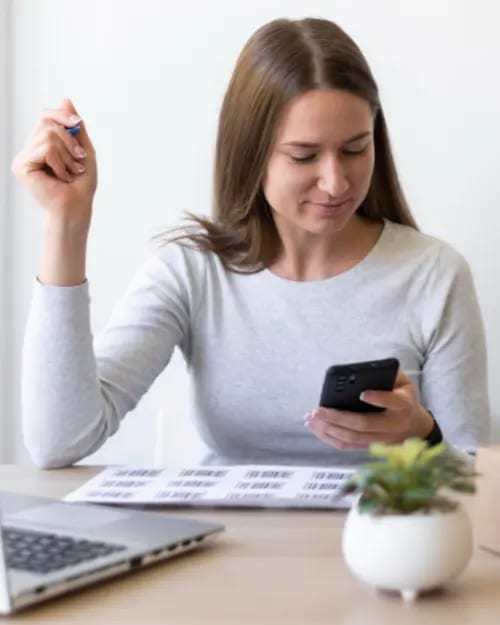 a person sits at a desk looking at a smartphone, holding a pen, with a laptop and papers on the desk, and a small potted plant in the foreground—deep in thought about mastering continuity programs to establish sustainable income streams.